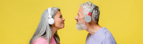 side view of funny multiethnic and middle aged couple in wireless headphones looking at each other isolated on yellow, banner