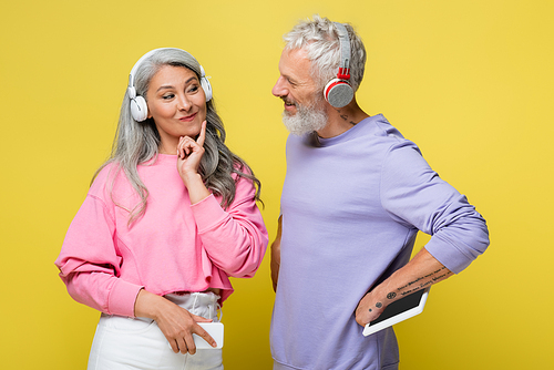 cheerful middle aged and multiethnic couple in wireless headphones looking at each other and holding gadgets isolated on yellow
