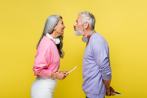 side view of funny multiethnic and middle aged couple in wireless headphones looking at each other and grimacing isolated on yellow