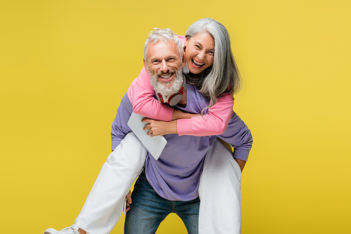 cheerful middle aged man piggybacking happy asian wife with digital tablet isolated on yellow