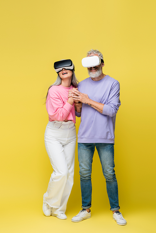 full length of multiethnic and middle aged couple in vr headsets holding hands while gaming on yellow