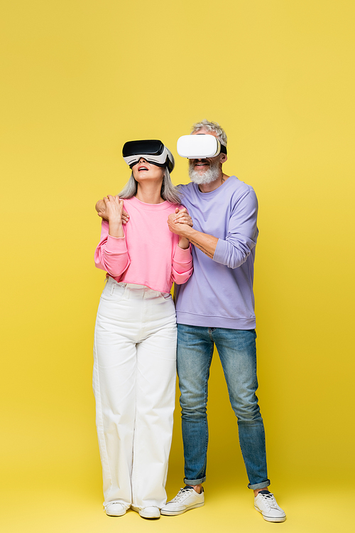 full length of interracial and middle aged couple in vr headsets holding hands while gaming on yellow