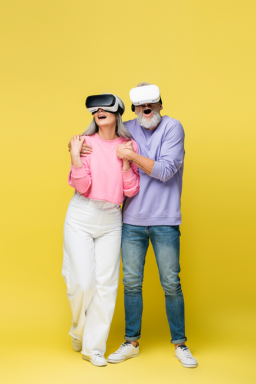 full length of interracial and surprised middle aged couple in vr headsets holding hands while gaming on yellow