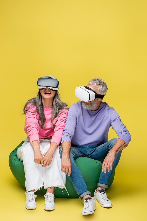 interracial and happy middle aged couple in vr headsets sitting in bean bag chair on yellow