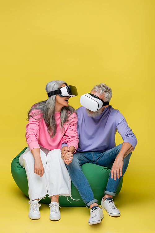 interracial and cheerful middle aged couple in vr headsets sitting in bean bag chair on yellow