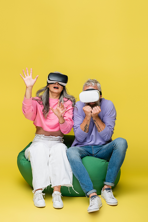 interracial and scared middle aged couple in vr headsets sitting in bean bag chair while gaming on yellow
