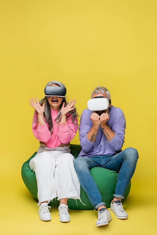 interracial and afraid middle aged couple in vr headsets sitting in bean bag chair while gaming on yellow