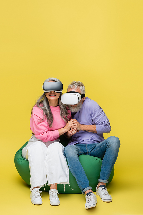 interracial and scared mature couple in vr headsets holding hands and sitting in bean bag chair while gaming on yellow