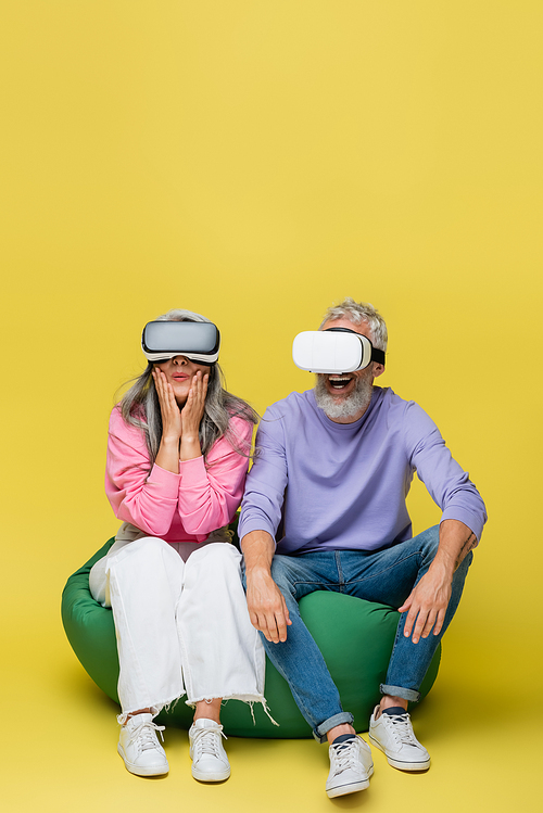 interracial middle aged couple in vr headsets sitting in bean bag chair while gaming on yellow
