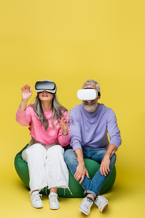 interracial and middle aged couple in vr headsets sitting in bean bag chair and gaming on yellow