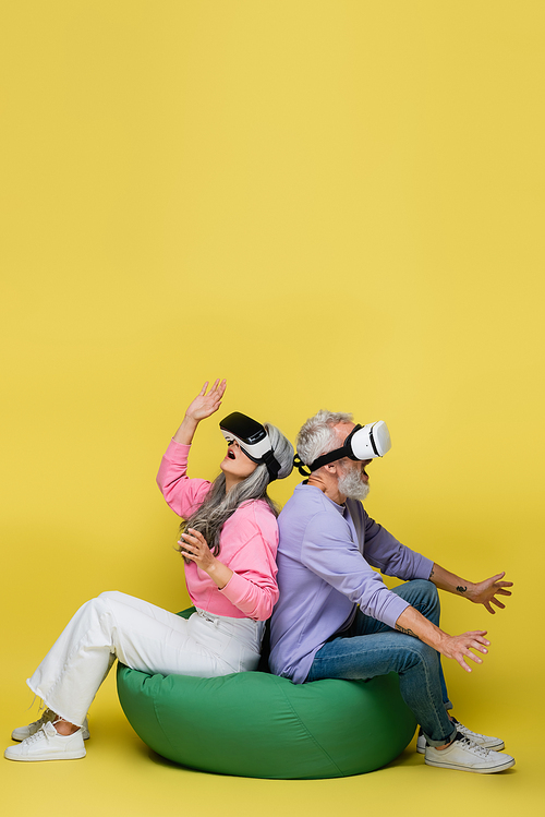 interracial and surprised middle aged couple in vr headsets sitting in bean bag chair while gaming on yellow