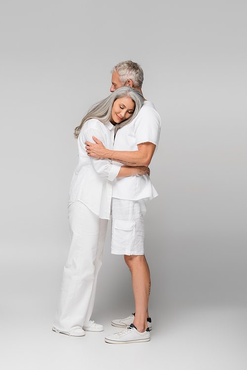 full length of happy interracial and mature couple hugging on grey