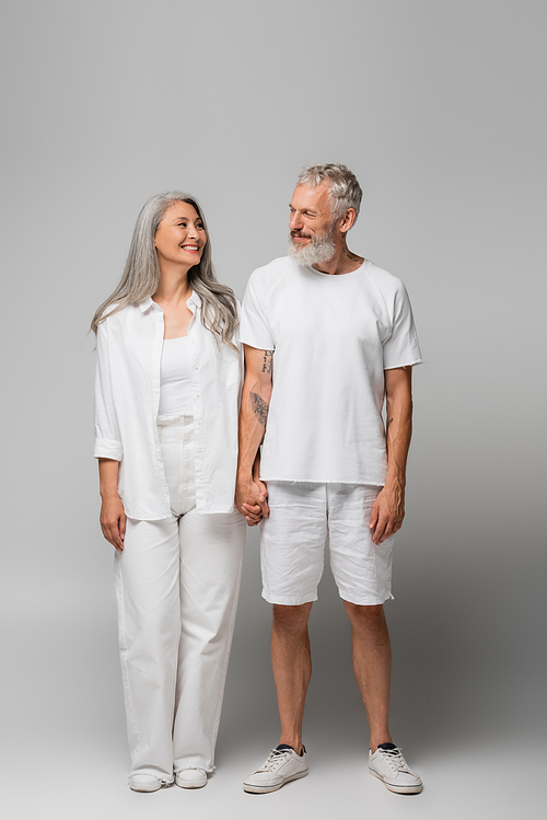 full length of happy interracial and mature couple holding hands on grey
