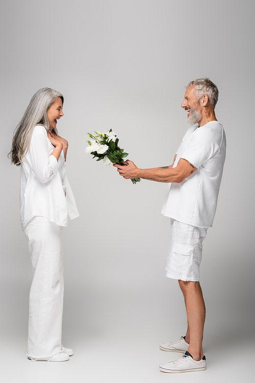 side view of happy middle aged man giving bouquet of flowers to amazed asian wife on grey