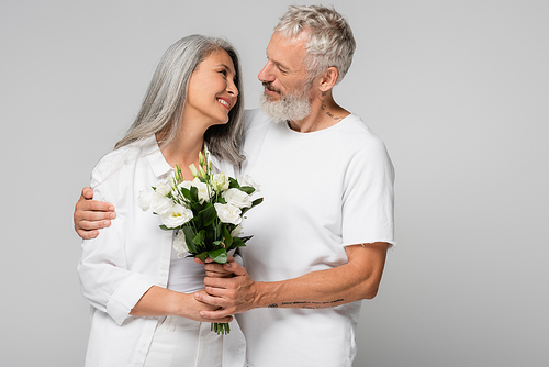 happy interracial and mature couple holding flowers and looking at each other isolated on grey