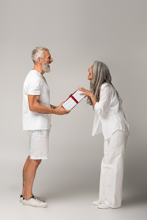 full length of cheerful middle aged man giving wrapped gift box to amazed asian wife on grey