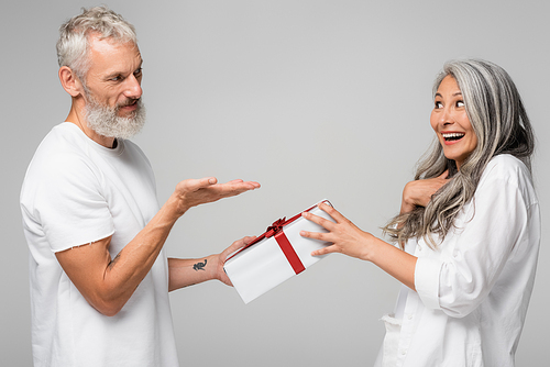 happy middle aged man pointing with hand at amazed asian wife while giving wrapped gift box isolated on grey