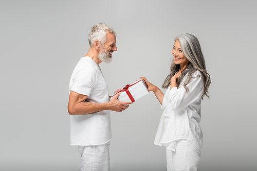 happy middle aged man giving wrapped gift box to smiling asian wife isolated on grey