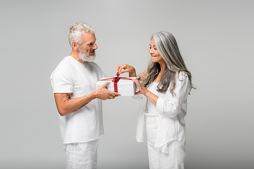 happy middle aged man holding wrapped gift box near curious asian wife isolated on grey