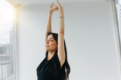 smiling armenian woman stretching with raised hands and closed eyes near window