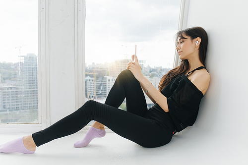 young armenian woman with smartphone listening music while sitting on floor near window