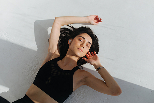 top view of pretty armenian woman in wireless earphone relaxing on white floor with shadows