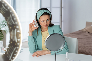 stylish woman proposing face serum near ring lamp and mirror