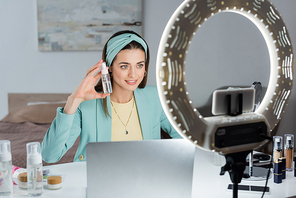 stylish woman showing spray bottle of micellar water near laptop and phone holder with ring lamp