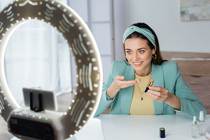 smiling woman pointing at lipstick near smartphone in holder with circle lamp