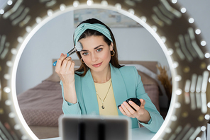 blurred smartphone in holder with circle light near beauty blogger applying face powder