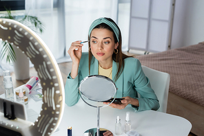 pretty woman applying eye shadows near mirror and smartphone in holder with ring light