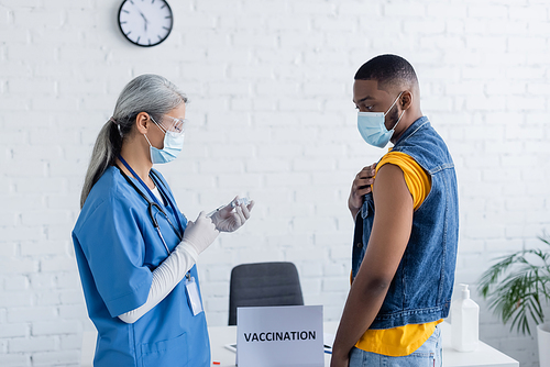 asian doctor in medical mask and goggles filling syringe with vaccine near african american man