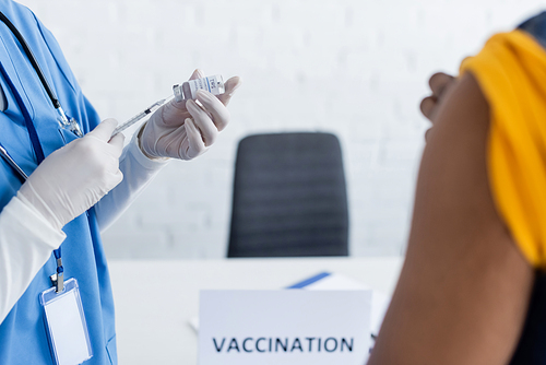 partial view of doctor in latex gloves holding vaccine near blurred african american man in vaccination center