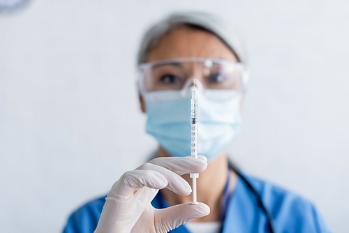 blurred asian immunologist in medical mask and goggles holding syringe with vaccine