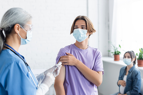 doctor in medical mask and latex gloves vaccinating young man near blurred pregnant woman