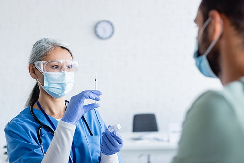 mature asian doctor in medical mask and goggles preparing syringe with vaccine near blurred man