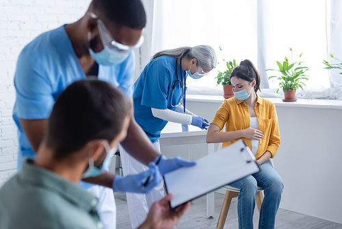 african american doctor in medical mask holding clipboard near blurred man in vaccination center