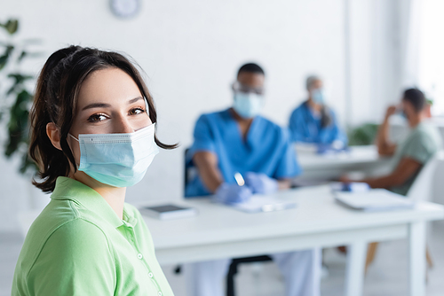 young woman in medical mask  near blurred multiethnic doctors in vaccination center