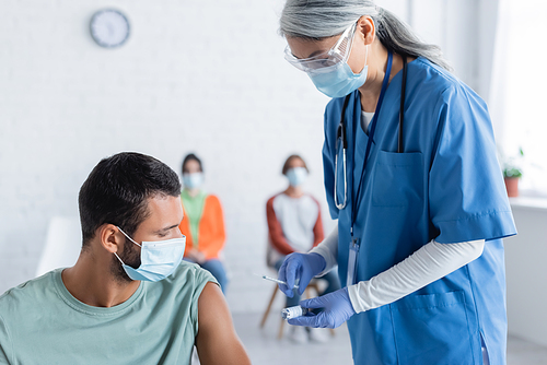 mature asian physician in medical mask holding syringe and jar with vaccine near young man and blurred patients