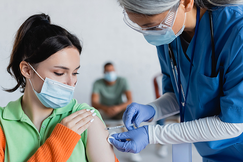 mature asian nurse in medical mask, goggles and latex gloves vaccinating young woman