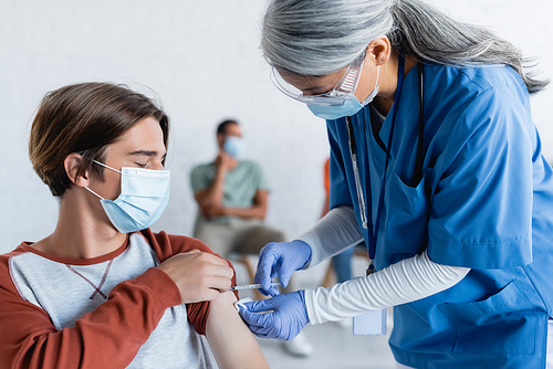 mature asian in medical mask vaccinating young patient near blurred african american man