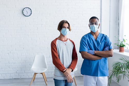 african american doctor standing with crossed arms near young man in medical mask, vaccination concept