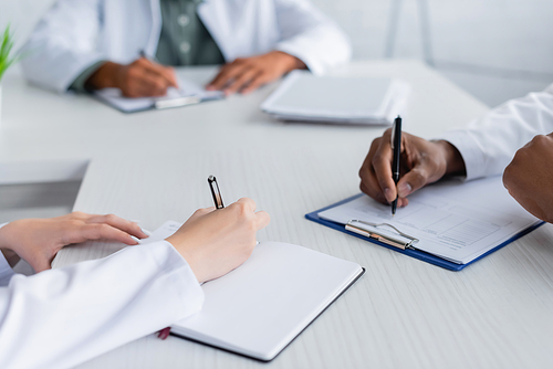 cropped view of multiethnic doctors writing during council in meeting room