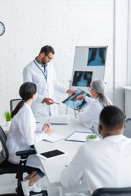 mature doctor pointing at lungs x-ray during council with multiethnic colleagues