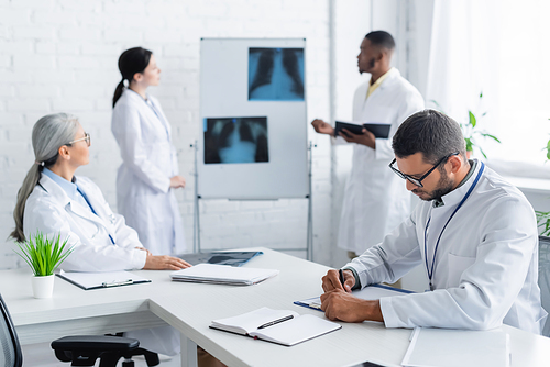 young doctor writing on clipboard while blurred multiethnic colleagues talking near lungs x-rays