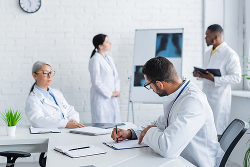 young doctor writing in notebook while multicultural colleagues talking near lungs x-rays on blurred background