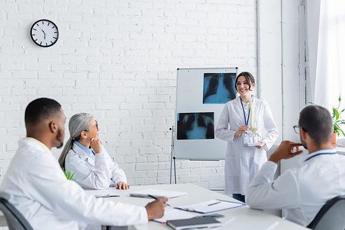 smiling doctor standing near flip chart with lungs x-rays and showing spine model to multiethnic colleagues