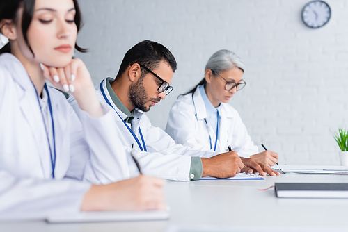 young physician in eyeglasses writing near multiethnic colleagues in meeting room