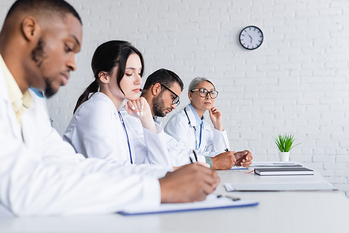 blurred african american doctor writing near colleagues during council in hospital