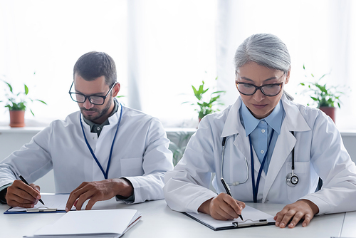 young and mature interracial doctors in eyeglasses writing on clipboards